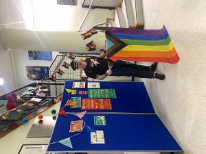 A person stands at a podium draped with a Progress Pride flag, speaking at a Pride event in the UCD Veterinary Medicine foyer. The space is decorated with rainbow flags and colourful bunting.